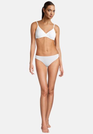 Giovane donna con capelli scuri che indossa un reggiseno bianco e mutandine abbinate, in piedi a piedi nudi contro uno sfondo chiaro e semplice.