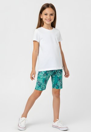 Młoda dziewczyna uśmiechająca się, ubrana w białą koszulkę, zielone kwieciste szorty za kolano oraz białe trampki, stojąca na jednej nodze na jednolitym tle.