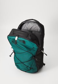 Mochila en verde azulado y negro con un zipper en la parte superior, cuerdas elásticas ajustables en la parte frontal y un forro interior gris. Cuenta con tirantes acolchados y bolsillos laterales.