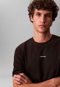 Must puuvillane t-särk ümmarguse kaelusega ja lühikeste varrukatega. Ees valge "Calvin Klein" logo. Sile tekstuur, keha lähedane lõige.