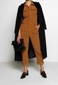 Mono de pana marrón con puños doblados, dos bolsillos en el pecho, llevado debajo de un abrigo largo negro. Bolso negro y zapatos de tacón bloque bicolor.