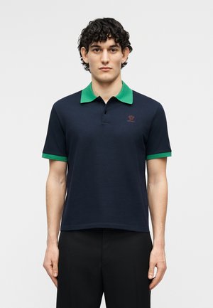 Νεαρός με σγουρά σκούρα μαλλιά φοράει μπλε πόλο μπλουζάκι με πράσινο γιακά και άκρες στα μανίκια, στέκεται μπροστά σε απλό φωτεινό φόντο.