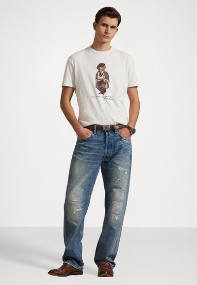 T-shirt blanc avec un imprimé graphique d'un ours en vêtements, associé à un jean bleu délavé avec des déchirures, et des chaussures en cuir marron.