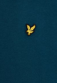 Tessuto a maglia turchese con finitura strutturata; presenta un piccolo logo di un'aquila gialla ricamata contornato di nero sul lato superiore sinistro.