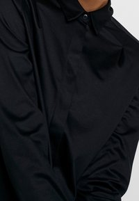 Chemise noire à manches longues avec un col à boutons, présentant un tissu lisse et une texture plissée le long des manches.