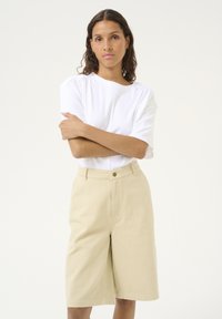Witte korte mouwtop met subtiele plooien op de schouders, gecombineerd met lichtbeige knielange shorts met een voorknoop en ceintuurlussen.