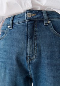 Close-up van de tailleband van een blauwe spijkerbroek met knoop en ritssluiting, gecombineerd met een ingestopt wit overhemd.