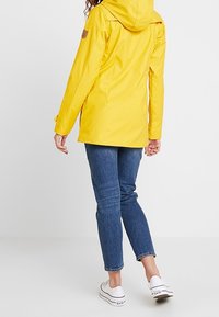 Gul vattentät jacka med huva, med en texturerad yta och ett logotypmärke på vänster ärm. Bärs över blå jeans och vita sneakers.