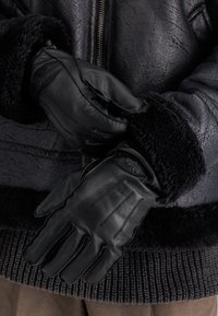 Zwarte leren handschoenen met een gladde textuur en schapenvacht manchet, met stiksels en een subtiel logo op één van de handschoenen.