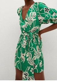 Robe fleurie verte avec un col en V, des manches bouffantes et une taille nouée. Présente des motifs botaniques blancs sur un tissu texturé.