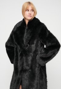 Svart fuskepelsjakke med vid krage, lange ermer og avslappet passform. Teksturen er myk, med et rikt, bustete utseende.