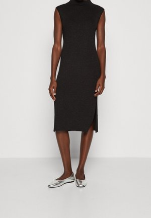 Femme portant une robe midi noire sans manches avec une fente latérale et des chaussures plates argentées, debout sur un fond clair uni.