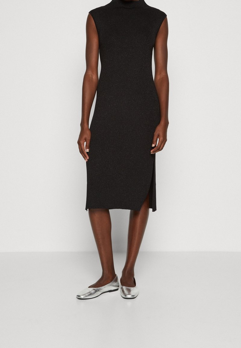 Femme portant une robe midi noire sans manches avec une fente latérale et des chaussures plates argentées, debout sur un fond clair uni.