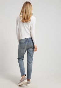 Bílý texturovaný svetr, světle modré zúžené džíny a šedé slip-on boty. Oblečení je vidět ze zadní strany, což zdůrazňuje jeho ležérní design.