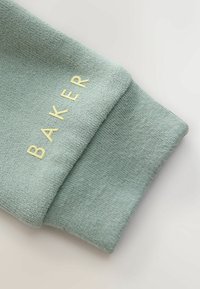 Jasnozielony mankiet swetra z teksturowanym materiałem dzianinowym. Na mankiecie znajduje się haftowane słowo "BAKER" w żółtym kolorze.