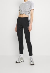 Šedé zkrácené tričko s bílým lemem, spárované s černými přiléhavými kalhotami s bílými postranními pruhy a logem adidas. Stříbrné sportovní boty.