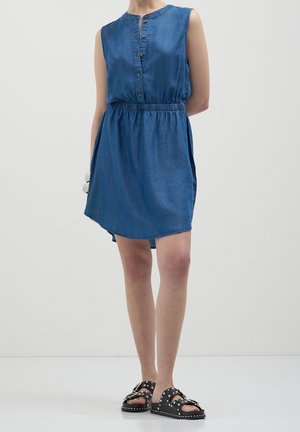 Donna che indossa un vestito di jeans blu senza maniche con bottoni frontali e sandali neri con borchie, in piedi su uno sfondo bianco semplice.