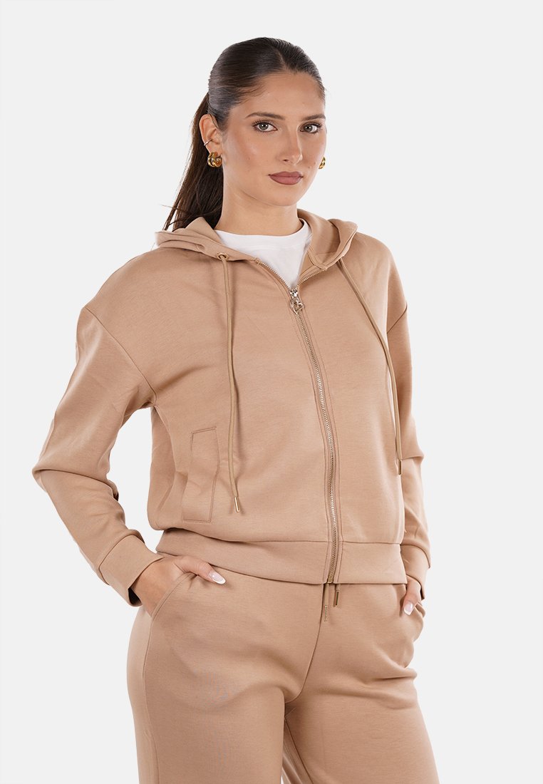 Donna con lunghi capelli scuri, in felpa beige con zip e pantaloni abbinati, mani in tasca, indossando orecchini a cerchio dorati, in piedi su uno sfondo chiaro.