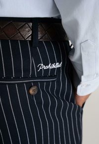 Close-up van donkerblauwe broek met streepjes, met "Verboden" geborduurd boven een knoopzak aan de achterkant en een bruine gestructureerde riem.