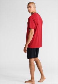 Rode t-shirt met korte mouwen en een stippenpatroon, ronde halslijn, voorzak, gecombineerd met donkerblauwe shorts. Model is iets zijwaarts gepositioneerd.