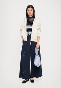 Young woman wearing cream zip hoodie, striped shirt, wide navy pants with text, holding a knotted light blue bag, standing on a white background.