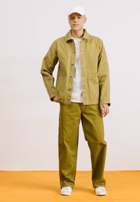 Chaqueta verde oliva con botones al frente, dos bolsillos y costuras blancas, combinada con pantalones verdes oliva a juego y zapatillas deportivas blancas.
