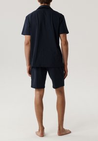 Osoba stojąca na boso, odwrócona tyłem, ubrana w granatową koszulę z krótkim rękawem i dopasowane szorty do kolan, na jednolitym tle.