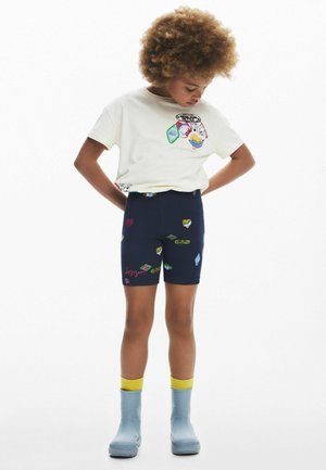 T-shirt crema chiaro con toppe grafiche colorate e pantaloni corti da bici blu navy con motivi multicolori di cuori e diamanti. Stivali da pioggia blu chiaro.