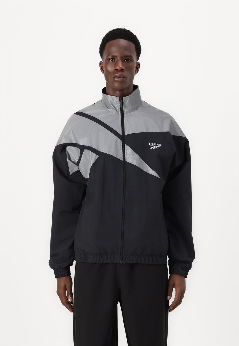 Schwarze und graue Windbreaker-Jacke mit hohem Kragen, Reißverschluss vorne und elastischen Bündchen. Mit diagonaler Farbblockierung und Branding.