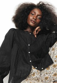 Blouse noire à manches longues en tissu texturé avec des accents froncés, dotée d'une fermeture à boutons sur le devant et d'un col. Pantalons à motif floral visibles.