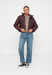 Doudoune bordeaux avec un col haut, à coupe courte et finition brillante, portée par-dessus un pull beige texturé et un jean bleu. Baskets noires.