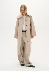 Beige trenčkot s velkým límcem a zapínáním na knoflíky, nošený přes bílé tričko a béžové kalhoty. Černé boty dokonalují celý outfit.