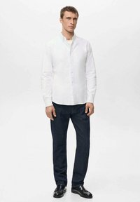 Camisa blanca abotonada con cuello mandarín, combinada con pantalones de mezclilla azul oscuro y zapatos negros. Tela lisa con un diseño ajustado.