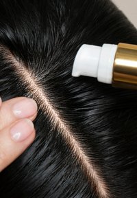 Cuir chevelu avec des cheveux noirs brillants séparés au milieu, montrant une main appliquant un embout blanc à pointe dorée distribuant un produit.
