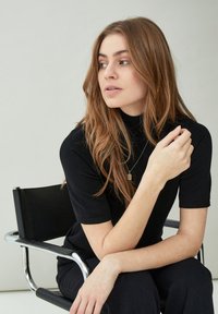 Young woman with long brown hair wearing black top and necklace, seated on black and chrome chair, looking to the side.