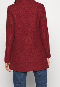 Rød uldfrakke med en tekstureret finish, der har en skræddersyet krave, lange ærmer og en let udvidet silhuet.