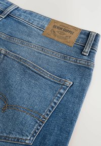 Plave traperice s kožnom etiketom na kojoj piše "DENIM SUPPLY", vidljivim šavovima i klasičnim dizajnom s pet džepova.