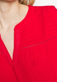 Blouse rouge avec un tissu texturé, dotée d'un décolleté en V et d'une bordure en dentelle. Des motifs subtils créent un intérêt visuel sur le haut.