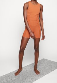 Personne debout pieds nus sur un tapis gris tacheté, portant une combinaison courte sans manches, ajustée, de couleur rouille avec des surpiqûres.