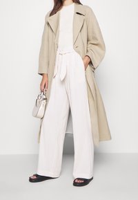 Trench-coat beige porté sur un haut blanc côtelé, associé à un pantalon blanc à jambes larges noué à la taille et des sandales noires, tenant un petit sac clair.