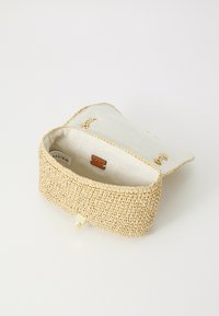 Geweven rieten tas in een tan kleur, met een stoffen klep bovenop, gouden hardware en een voering aan de binnenkant. De vorm is rechthoekig met een gestructureerde buitenkant.