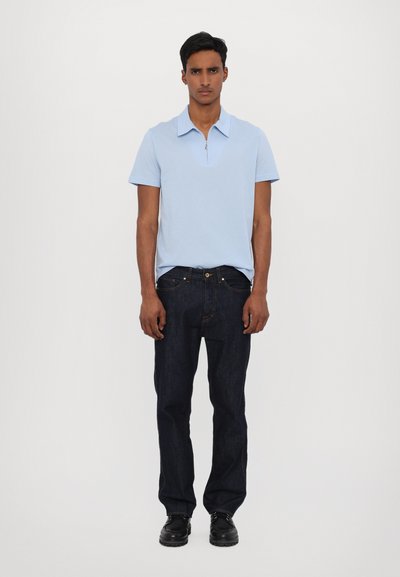 Ανοιχτό μπλε κοντομάνικο πόλο με φερμουάρ στο γιακά, συνδυασμένο με σκούρα μπλε τζιν και μαύρα παπούτσια, παρουσιασμένο σε όρθια μπροστινή θέση.