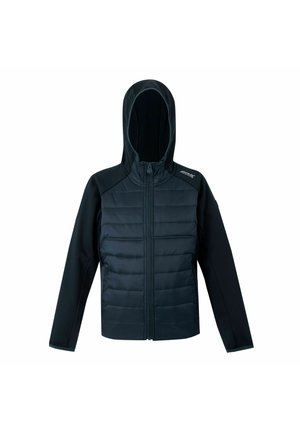 Junge steht mit den Händen in den Taschen, trägt eine dunkelblaue Jacke, passende Hose und schwarze sowie neon-grüne Sportschuhe.