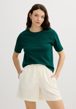 Mujer joven con cabello largo y castaño, vistiendo una camiseta de manga corta verde oscuro y pantalones cortos blancos de talle alto, de pie con las manos en los bolsillos.