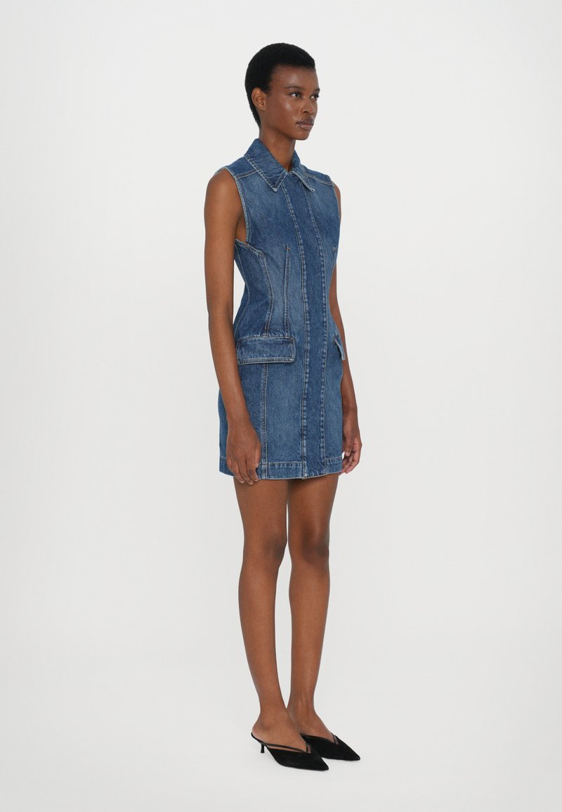 Ärmelloses Jeanskleid, hellblau, mit Kragen, vorne mit Reißverschluss, zwei vordere Taschen und taillierter Silhouette. Mit schwarzen Mules getragen.