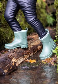 Bottes de pluie en caoutchouc vertes avec une semelle texturée, de hauteur cheville et doublées en polaire, placées sur une bûche au-dessus de l'eau.