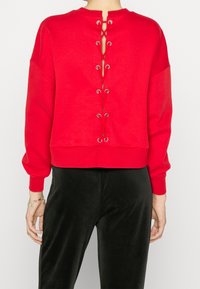 Sudadera roja corta con cuello redondo, mangas largas y cierre de cordones en la espalda con ojales metálicos. Combinada con pantalones negros.