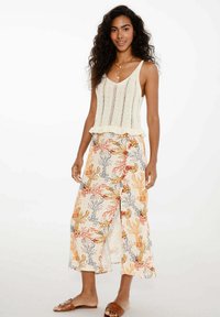 Pletený top bez rukávů v off-white barvě s vlnitým lemem, spárovaný s volnými, vzorovanými kalhotami se širokými nohavicemi, které mají květinové vzory v oranžové, šedé a žluté barvě.