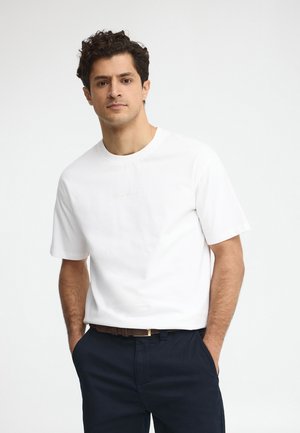 Man met donker krullend haar, een wit shirt met korte mouwen en een donkere broek, staand met handen in de zakken tegen een effen achtergrond.