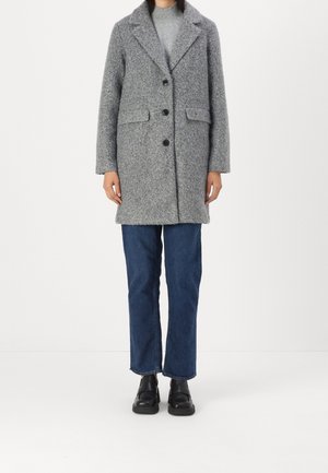 Personne portant un manteau en laine gris avec des boutons noirs, un jean bleu, des chaussures noires et un pull à col roulé gris clair, debout sur un fond blanc.